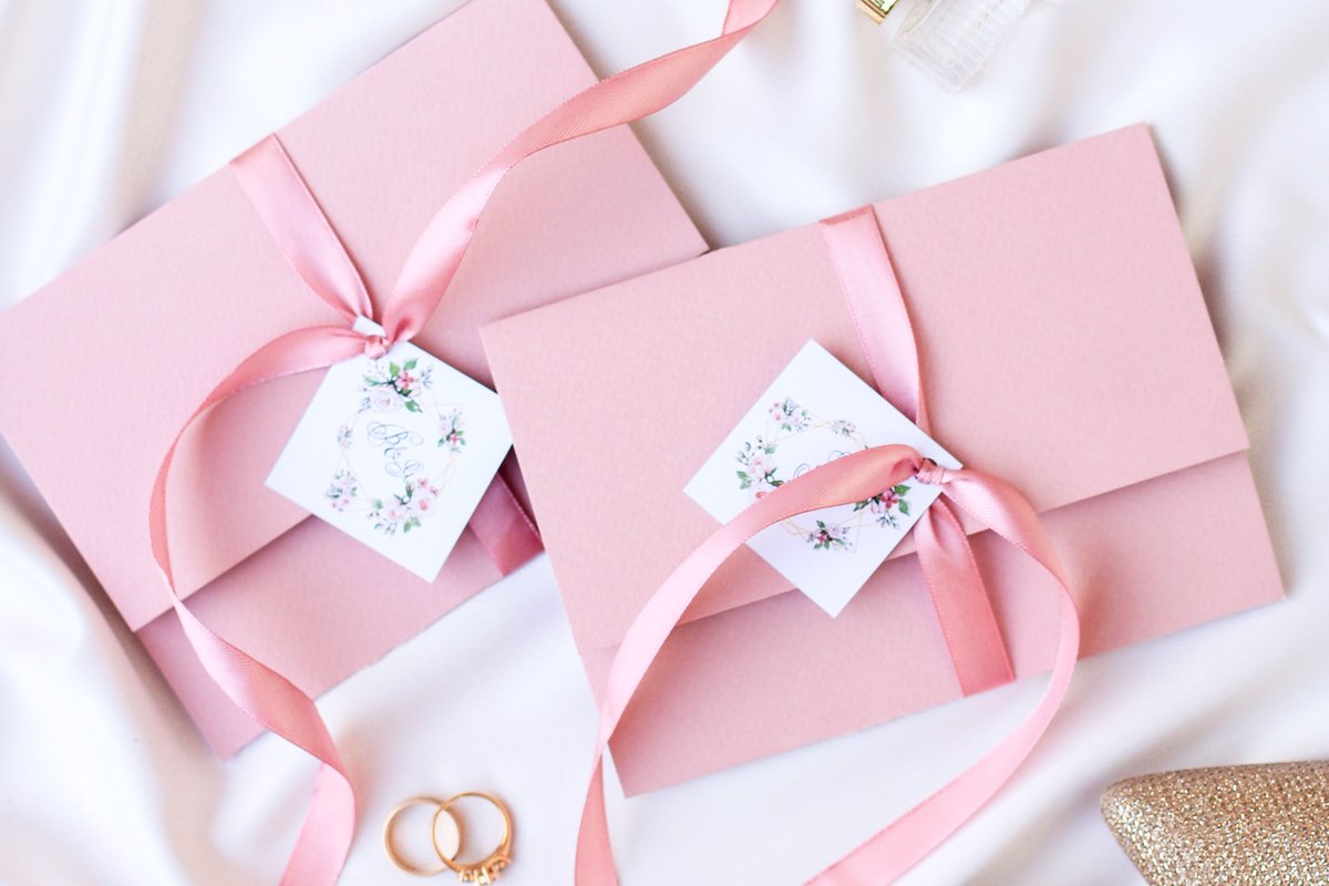 Blush pink wedding invitation envelopes tied with satin ribbon and monogrammed tags, holding the enclosure and RSVP cards that accompany an invitation